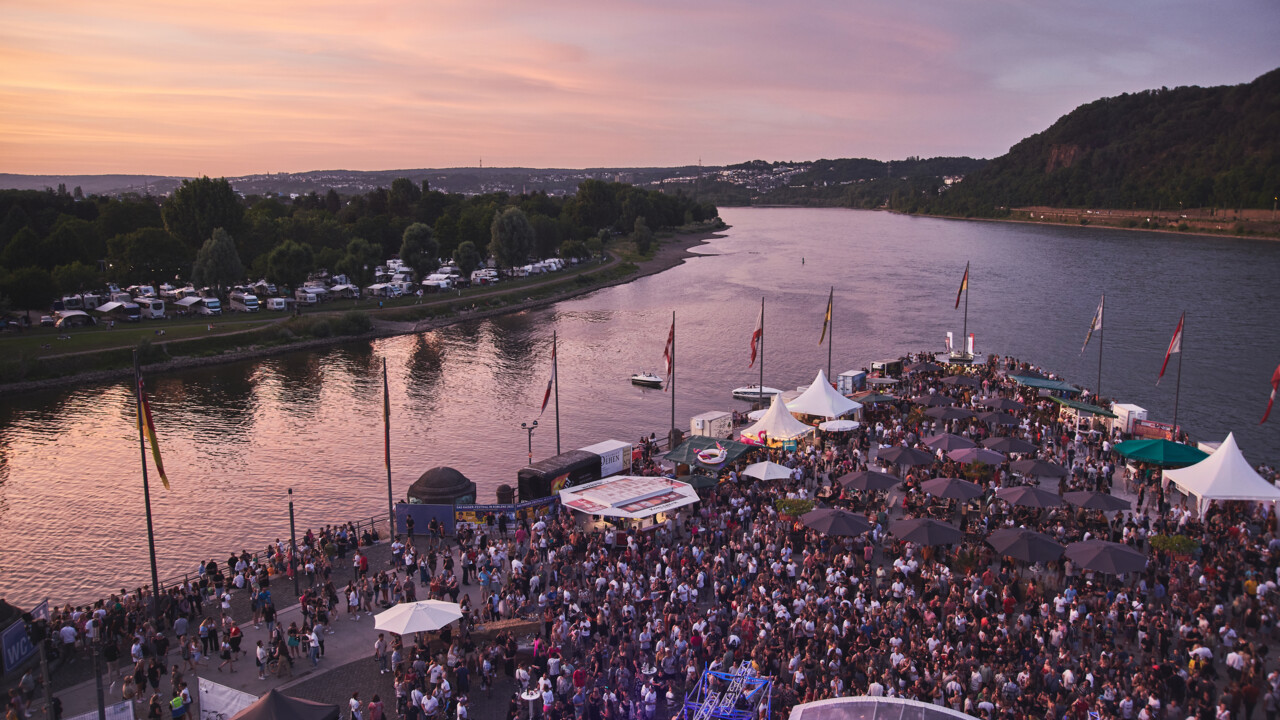 Blick von oben auf das Deutsche Eck bei Abenddämmerung während einer Veranstaltung, Menschengruppen stehen vor einer Bühne und an Ständen verteilt auf dem Gelände © Picture Colada
