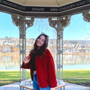 Frau mit rotem Mantel und Sonnenbrille steht im Augusta-Pavillon in den Rheinanlagen, im Hintergrund der Rhein und der Stadtteil Pfaffendorf. © Kaya Reichert