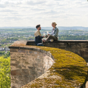 Pärchen sitzt auf der Mauer der Festung Ehrenbreitstein und hält Weingläser mit Stadt Koblenz im Hintergrund © Koblenz-Touristik GmbH, Dominik Ketz